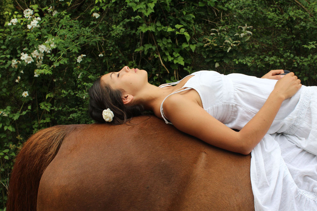 jeune femme sur la croupe de son cheval avec une robe blanche et une fleur dans les cheveux